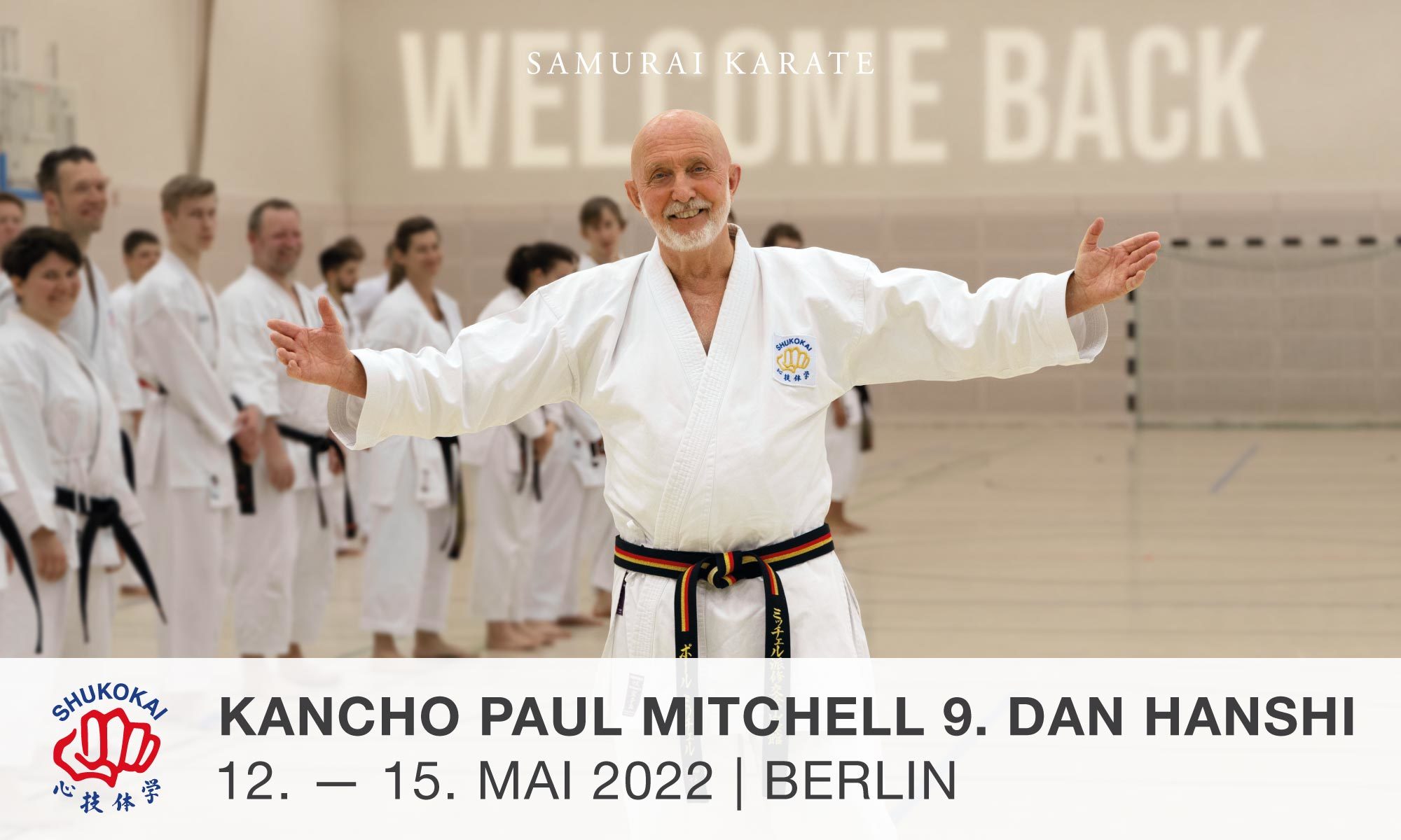 Kancho Paul Mitchell Hanshi steht im Karate-Anzug mit weit ausgebreiteten Armen für einer Gruppe Karate-Ka in einer Sporthalle.